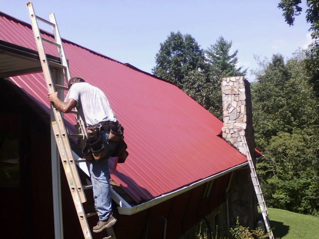 Roofer on a ladder beside a red metal roof and stone chimney — Soffit & Fascia Repair by Roofing Brothers in Glen Arm